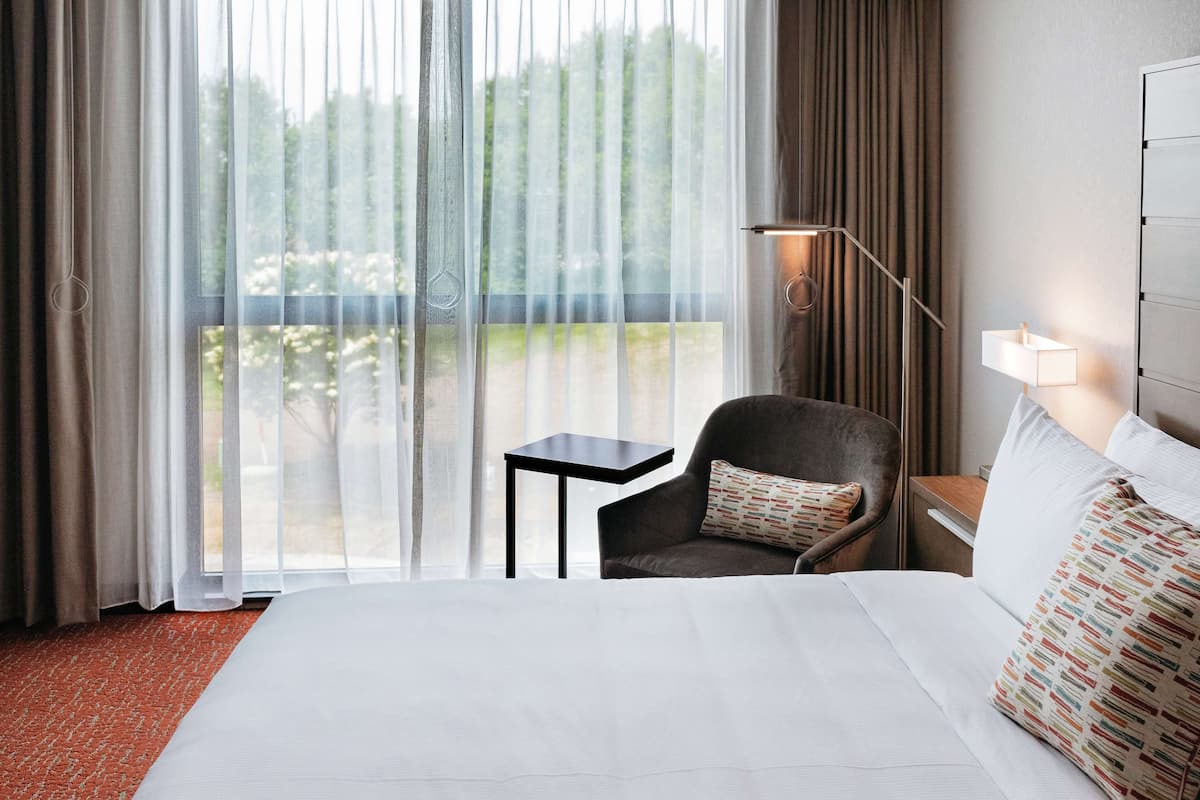Ein Hotelzimmer mit einem großen Bett, einem kleinen Tisch, einem Sessel und Blick auf Grün hinter dem Fenster.