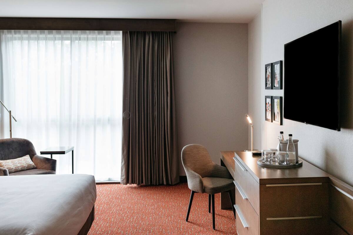 Ein Hotelzimmer mit Bett, Schreibtisch mit Tablett, Stuhl, Fernseher und einem Fenster mit Vorhängen.