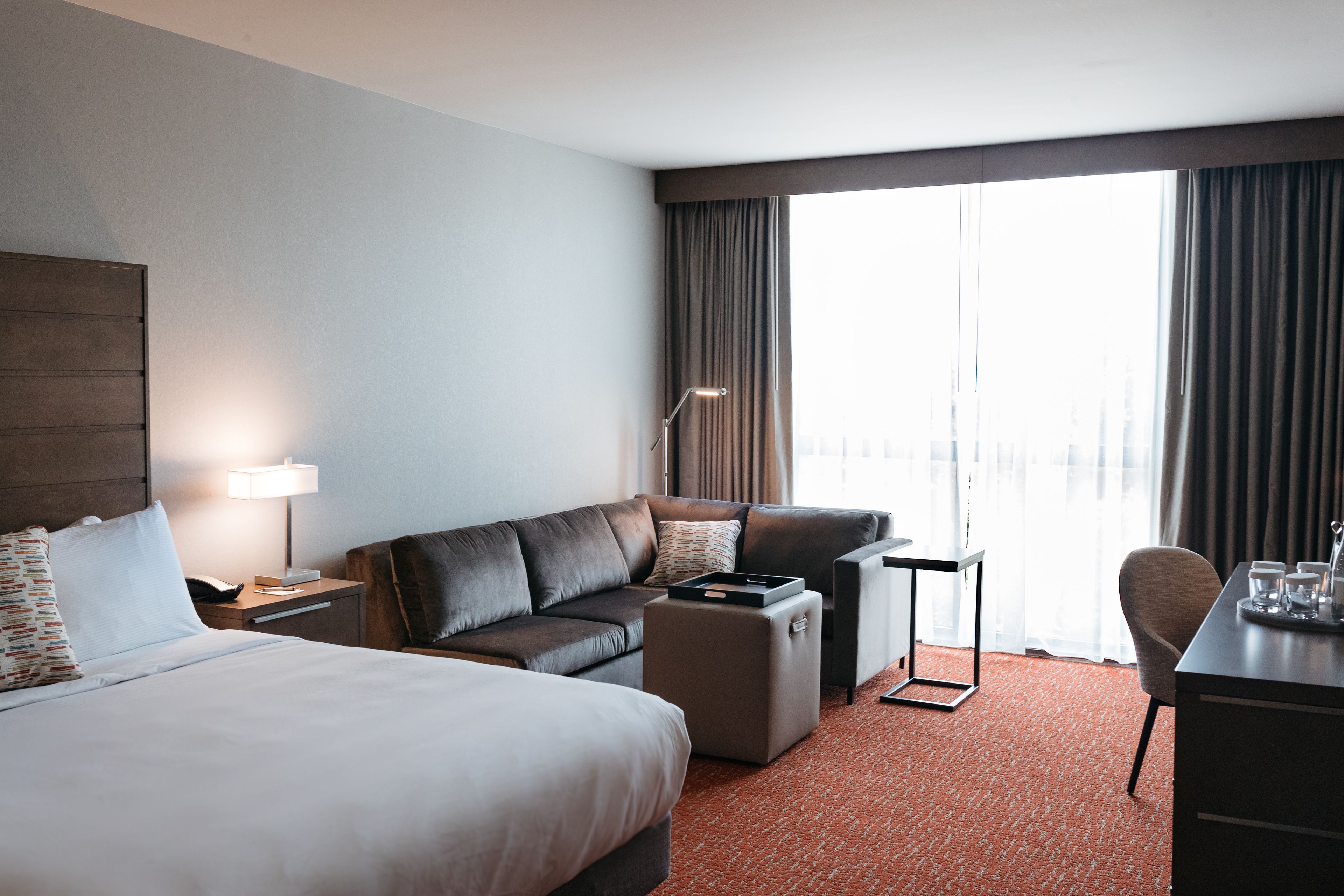 Habitación de hotel con una cama grande, un sofá, un escritorio y una silla. Hay una ventana con cortinas, una lámpara y una bandeja con vasos y una taza.