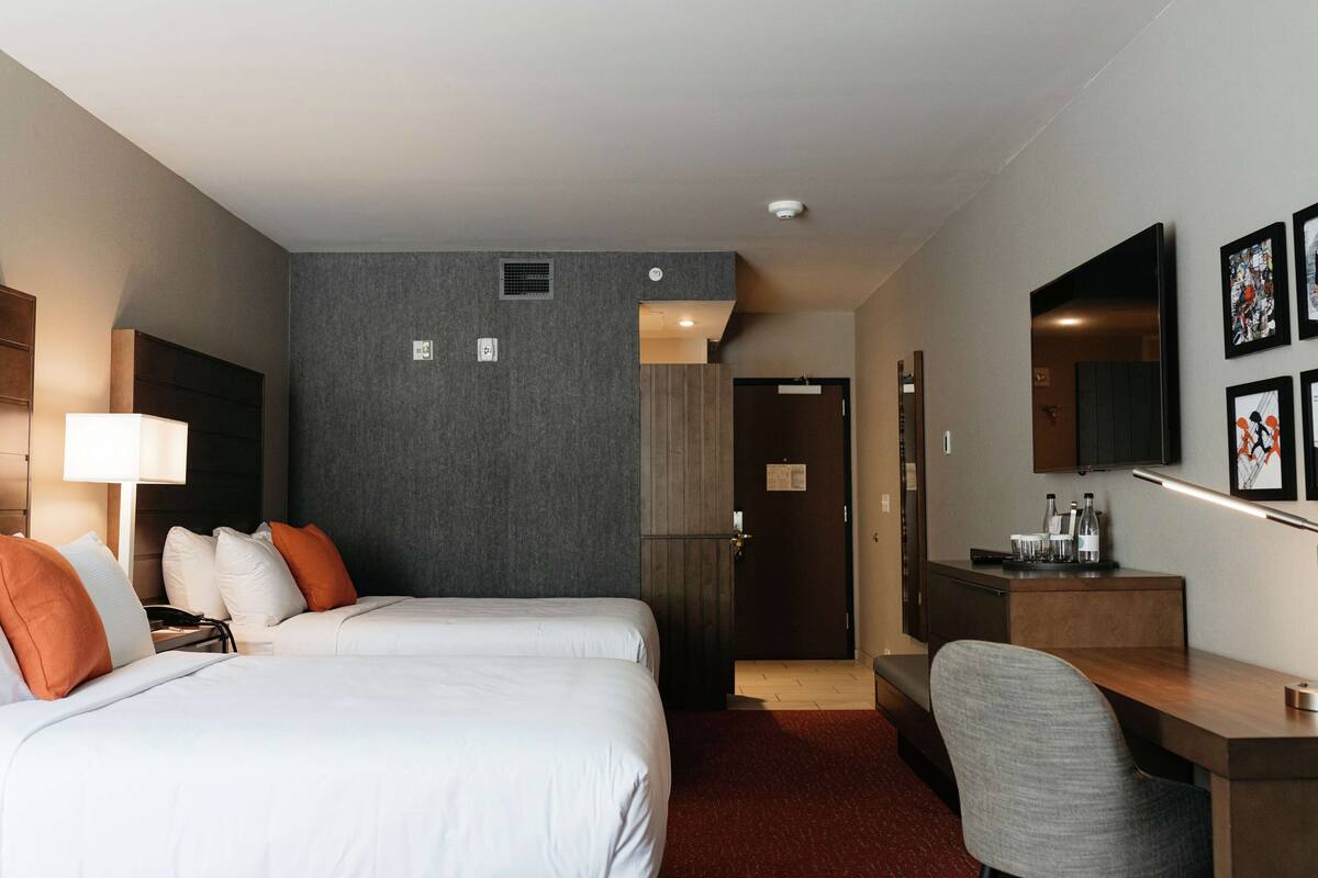 Ein Hotelzimmer mit zwei Betten, einem Schreibtisch, einem Stuhl, einer Lampe, einem Fernseher und Bildern an der Wand.