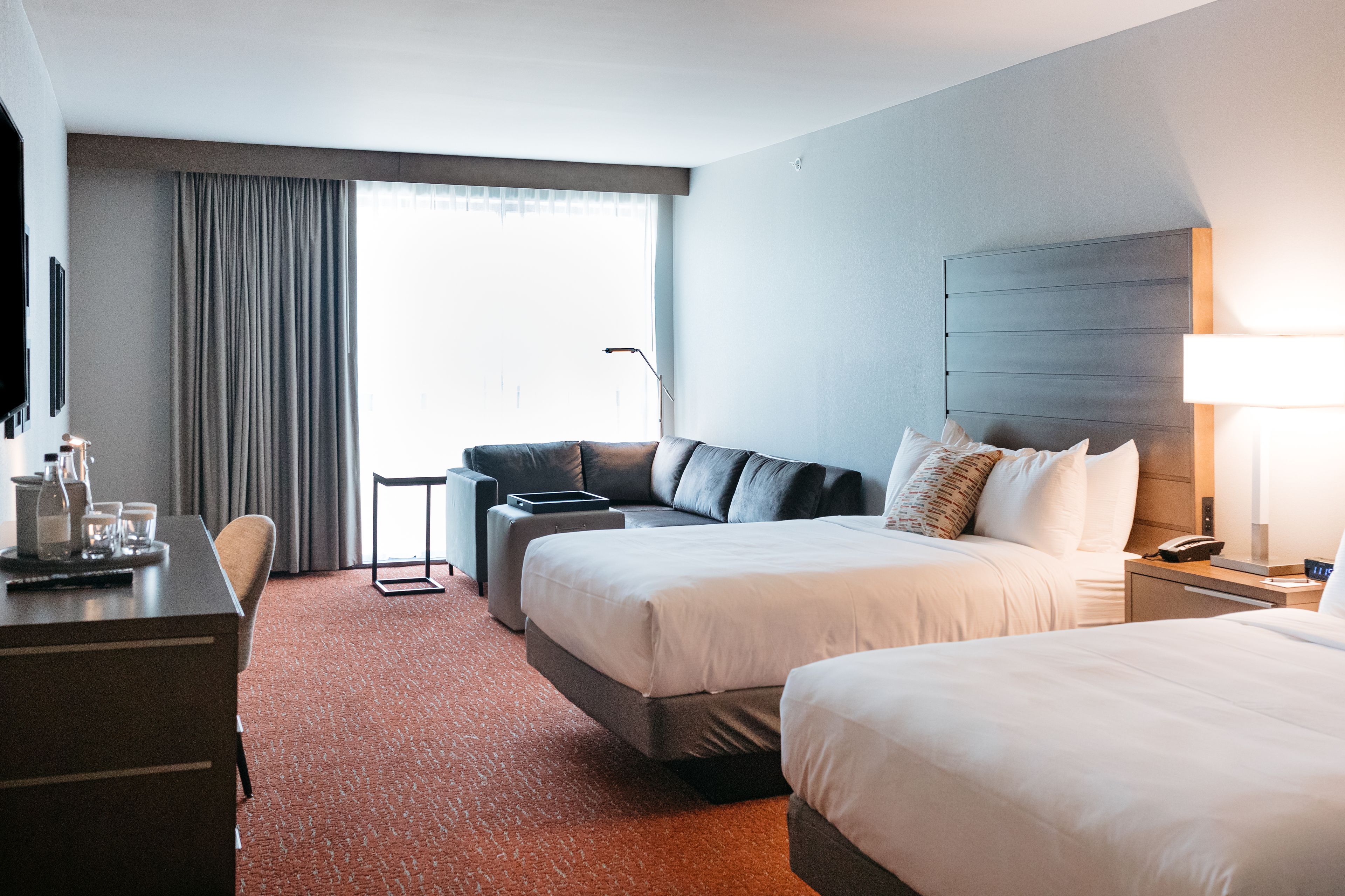 A hotel room with two beds, a desk, a chair, a sofa, and a large window with curtains.