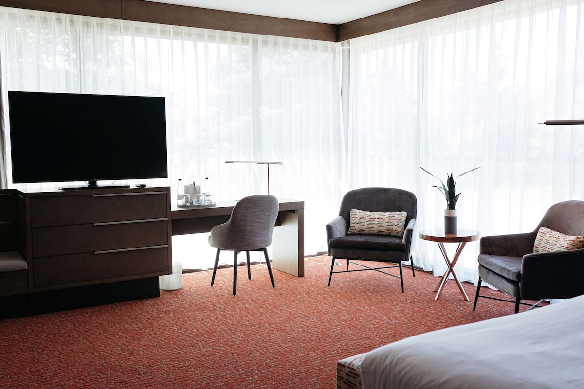 Ein Hotelzimmer mit einem großen Flachbildfernseher, einem Schreibtisch, zwei Sesseln, einem kleinen Beistelltisch mit Pflanze und einem Bett.