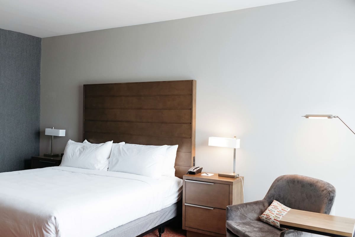 Ein Hotelzimmer mit einem großen Bett, einem Nachttisch mit Lampe, einem Sessel und einem Schreibtisch ebenfalls mit Lampe.