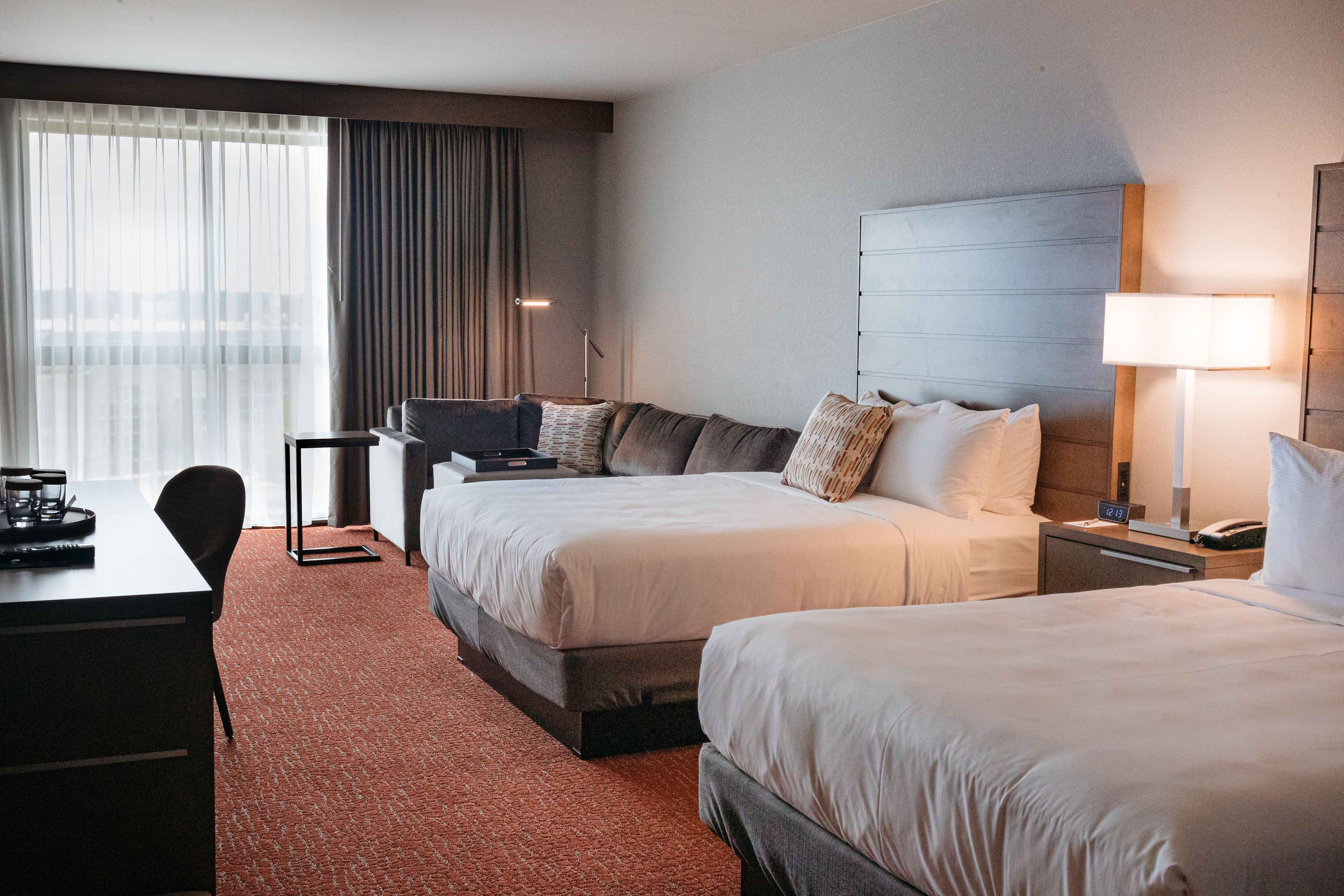 A hotel room with two beds, a desk, a chair, a lamp, and a view of the city through large windows.