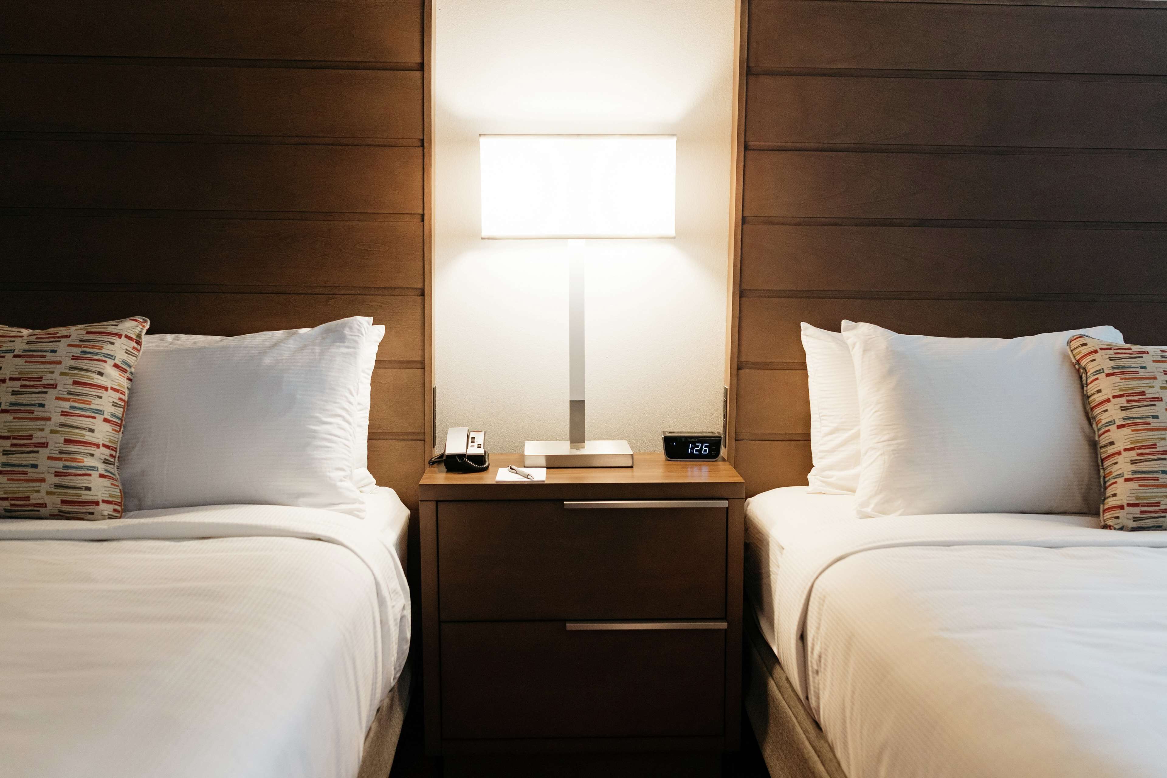 Habitación de hotel con dos camas, una mesita de noche con lámpara, teléfono y reloj digital.