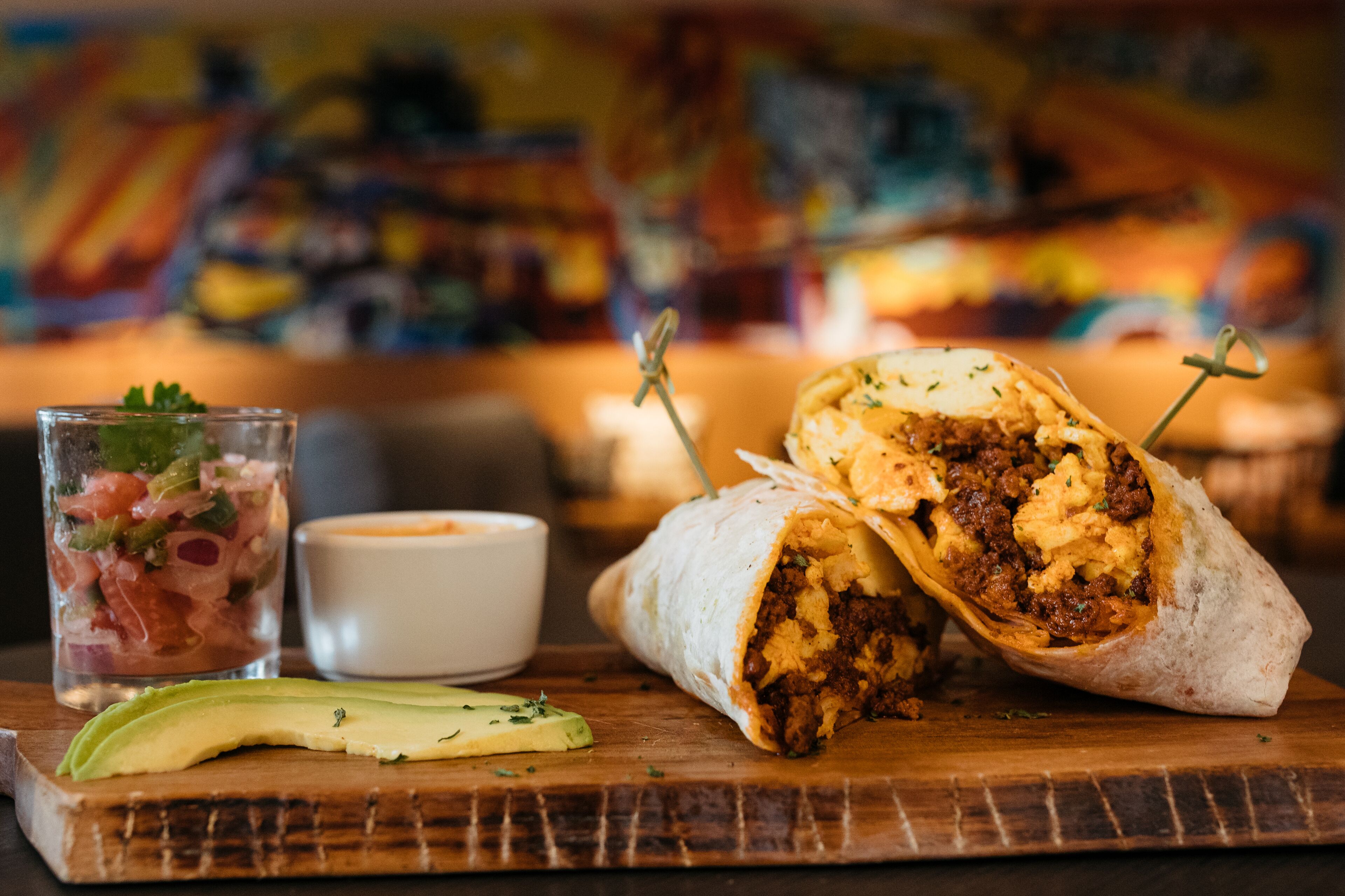 A burrito with a side of avocado and a small cup of sauce on a wooden board.
