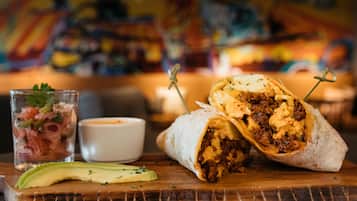 A burrito with a side of avocado and a small cup of sauce on a wooden board.