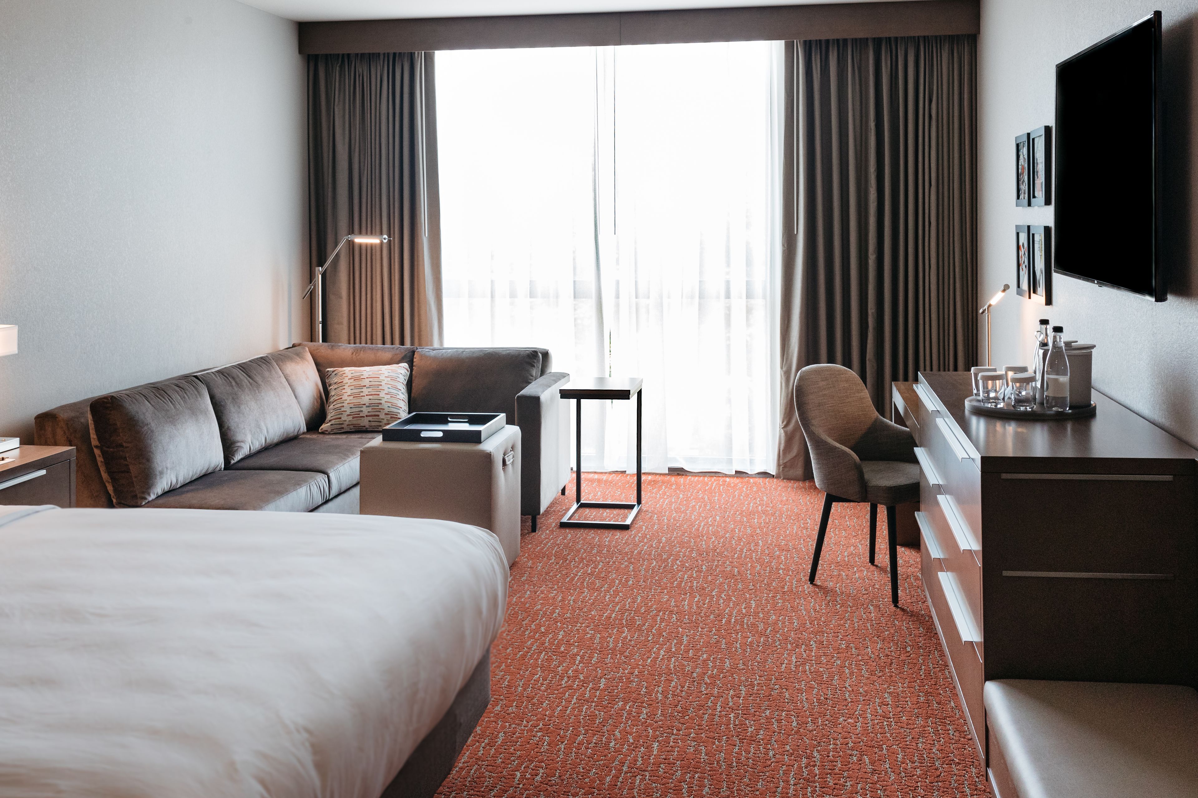 A hotel room with a bed, a sofa, a desk, and a chair. There is a large window with curtains, a flat-screen TV mounted on the wall, and a carpeted floor.