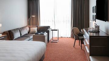 A hotel room with a bed, a sofa, a desk, and a chair. There is a large window with curtains, a flat-screen TV mounted on the wall, and a carpeted floor.