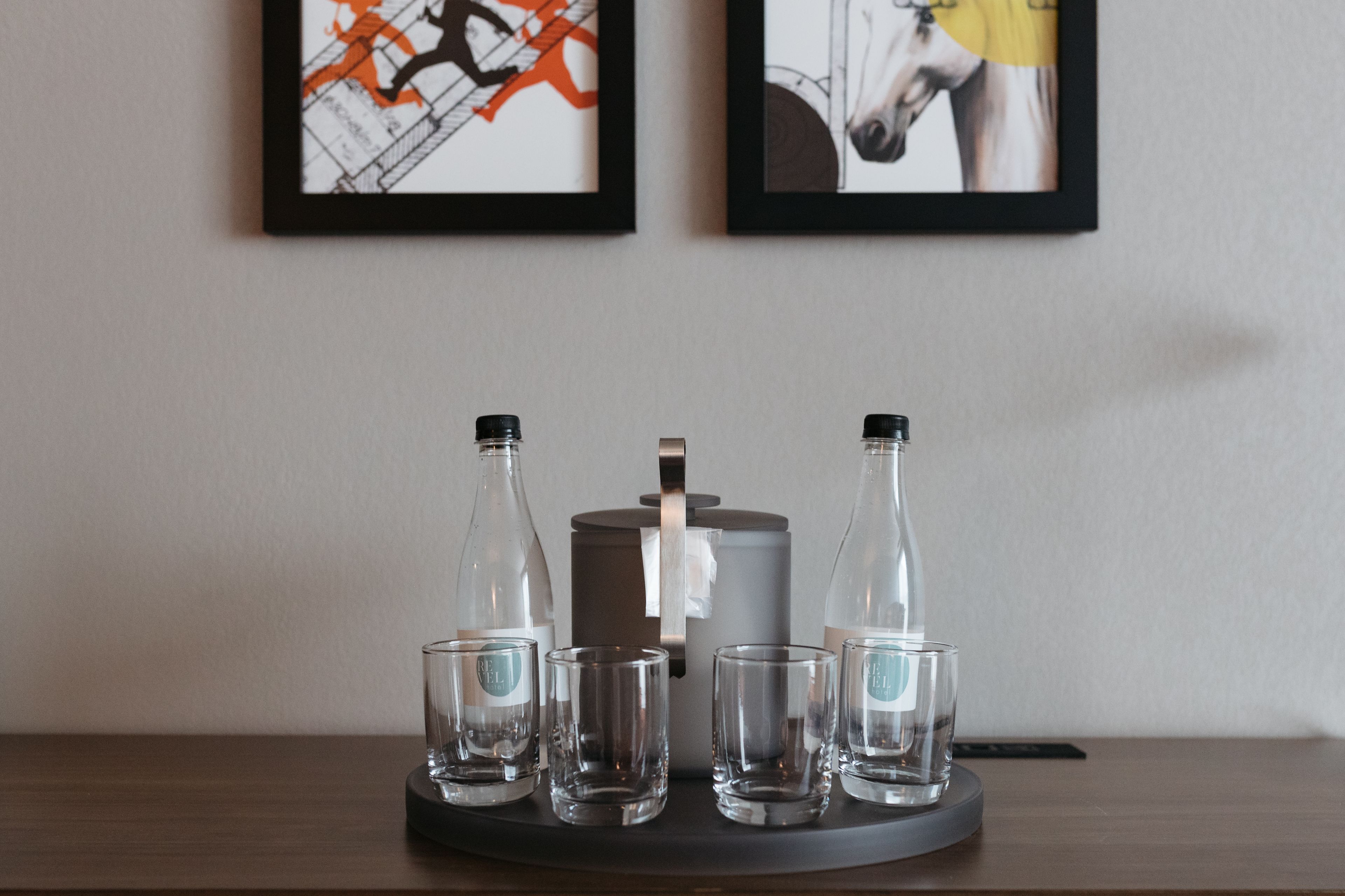 A table with four clear glass bottles and four empty glasses, a gray cylindrical object, and two framed artworks on the wall.