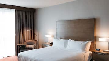 A hotel room with a large bed, a chair, a desk with a lamp, and a nightstand with a phone and clock.