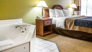 A hotel room with a bed, two bedside tables with lamps, a wooden nightstand, a bathroom with a bathtub and sink, and a window with curtains. - Quality Inn Darien - North Brunswick (Darien)