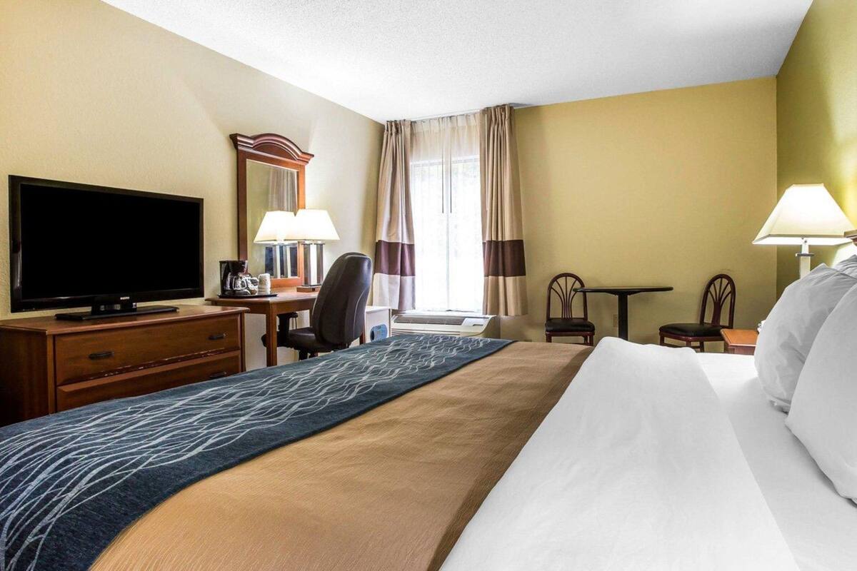 A hotel room with a large bed, a flat-screen TV, a desk with a chair, a small table, and a lamp.