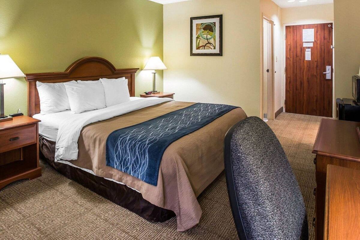 A hotel room with a bed, bedside lamps, a desk, a chair, a door, and a framed picture on the wall.