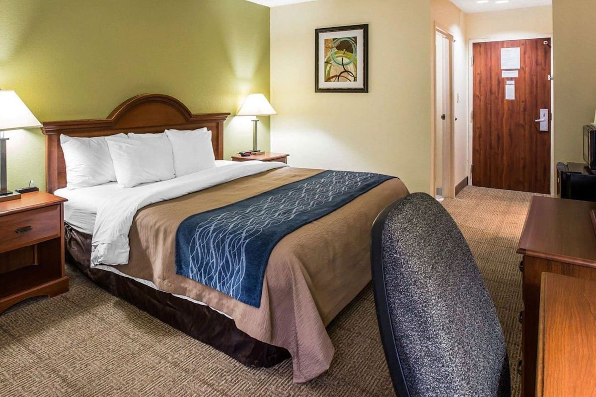 A hotel room with a bed, bedside lamps, a desk, a chair, a door, and a framed picture on the wall.