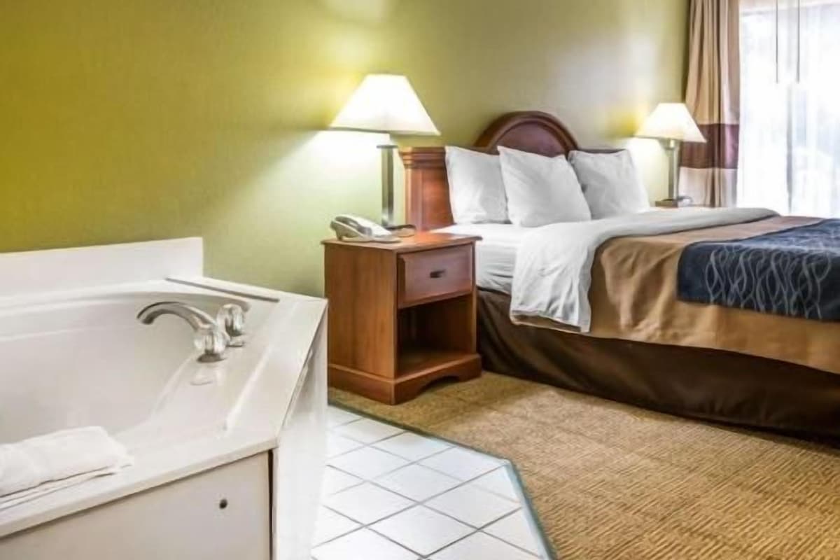Habitación de hotel con cama, dos lámparas de noche, mesita de noche, cabecero de madera, baño con bañera y suelo de baldosas.