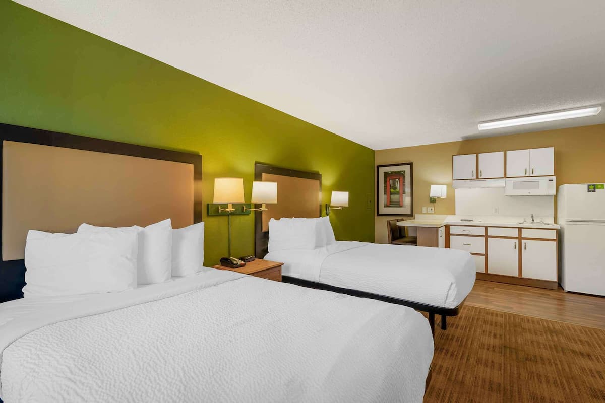 A hotel room with two beds, a kitchenette, and a small desk.