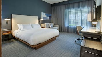 A hotel room with a large bed, a desk with a chair, a lamp, and a view of the outdoors through a window.