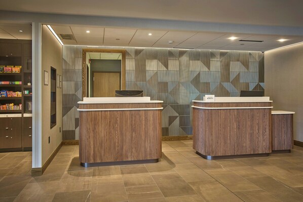 A reception area with two wooden reception desks, a wall with geometric patterns, and a shelf with snacks. - Courtyard by Marriott Wausau (Wausau)