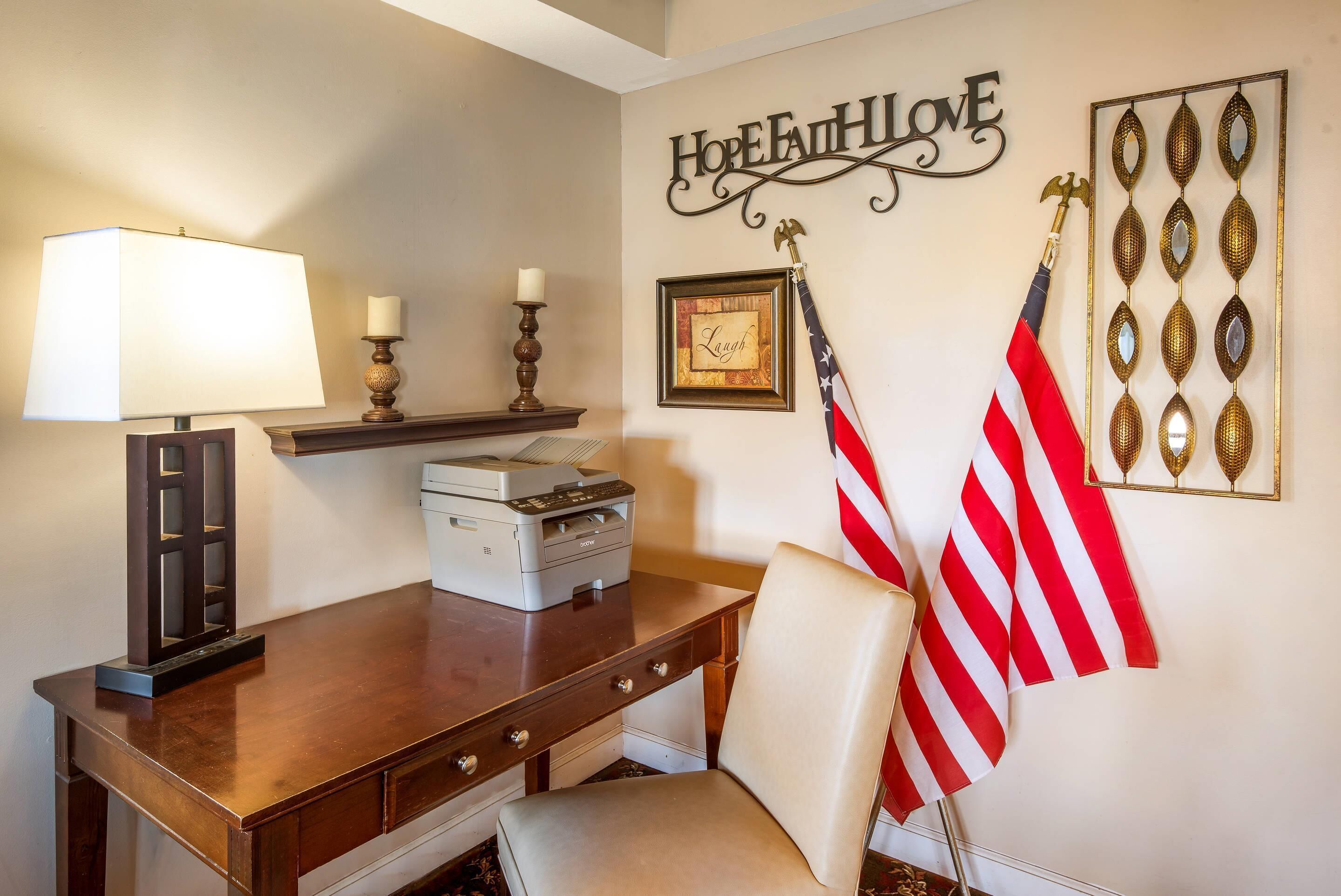 Una habitación con un escritorio de madera, una lámpara, una impresora, una bandera estadounidense, un cuadro enmarcado y un adorno de pared que dice “HOPE & LOVE”.