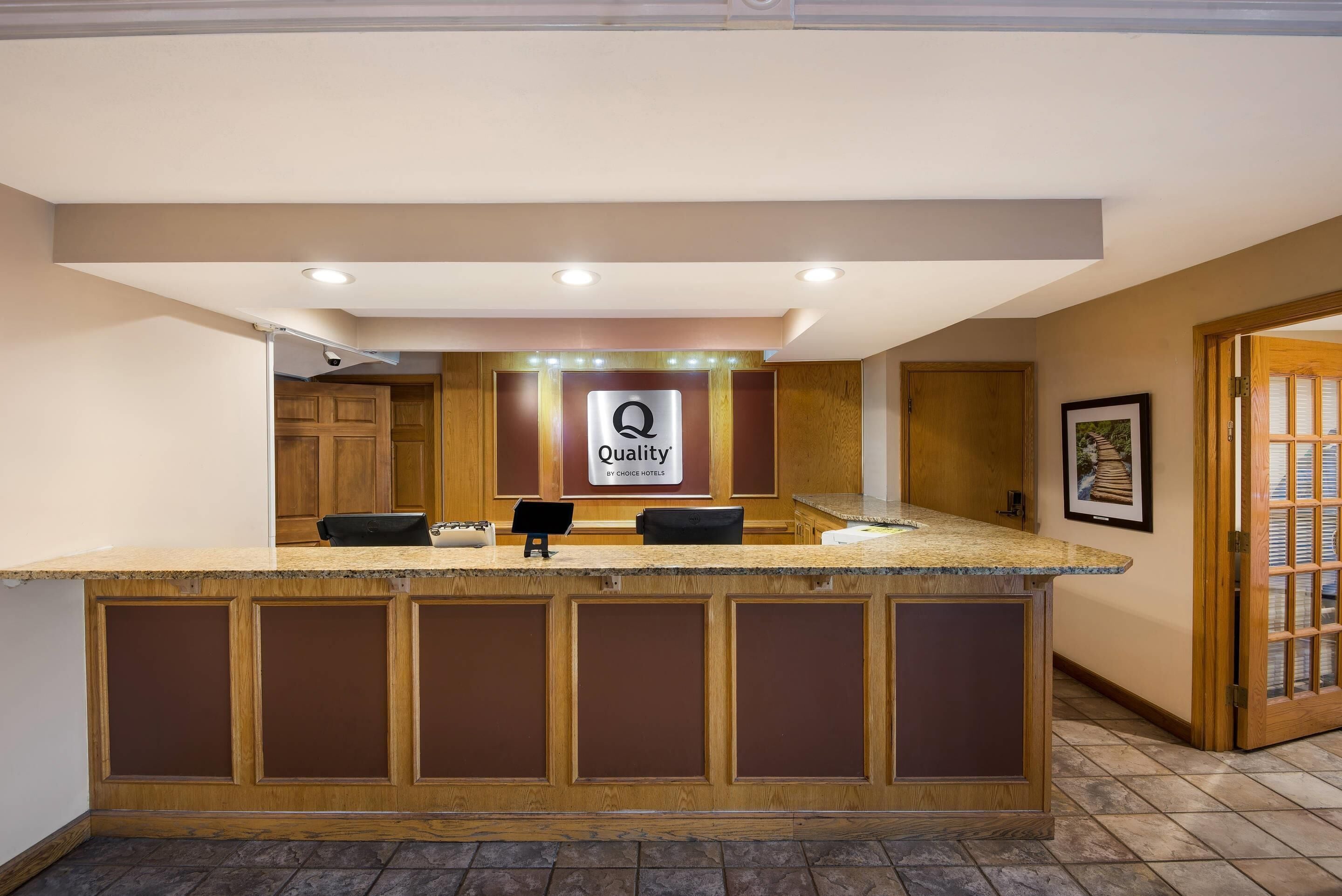 A hotel reception area with a wooden reception desk, a sign reading "Quality by Choice Hotels," and a framed picture on the wall.