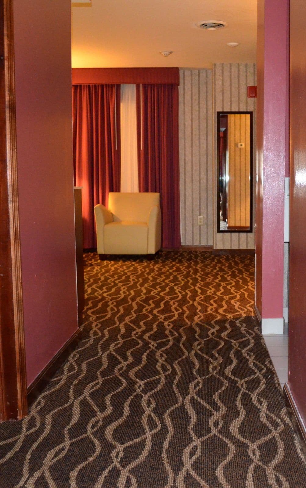 A hotel room with a patterned carpet, a cream-colored armchair, and a mirror on the wall.