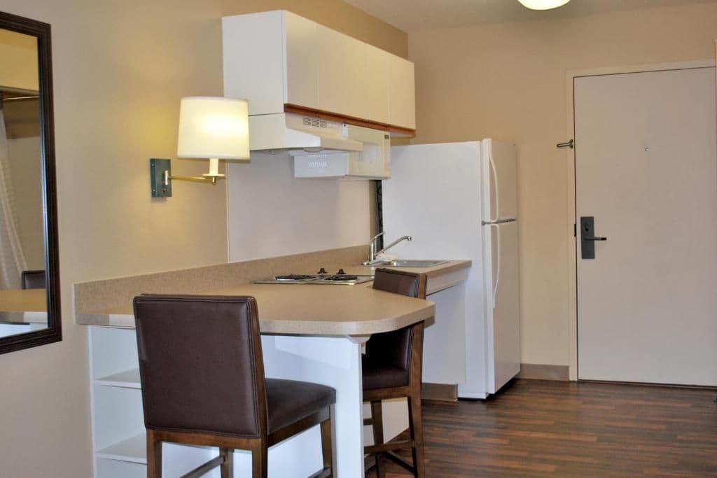 A compact kitchen with a countertop, two bar stools, a refrigerator, and a wall-mounted light fixture.