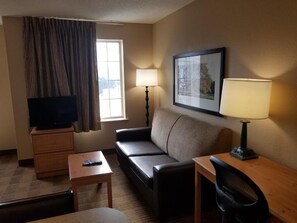 A hotel room with a sofa, a desk with a chair, a television on a stand, two lamps, and a framed picture on the wall. - Studio 6 Extended Stay Milwaukee Brookfield WI (Brookfield)