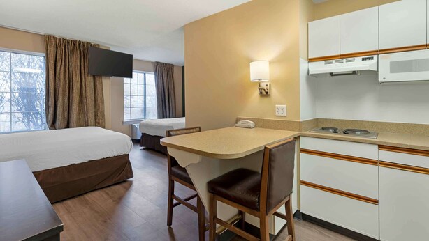 A hotel room with a bed, a desk with a chair, a kitchenette, a TV mounted on the wall, and a window with curtains.