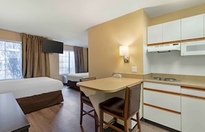 A hotel room with a bed, a desk with a chair, a kitchenette, a TV mounted on the wall, and a window with curtains.