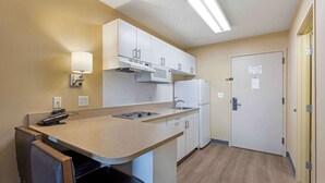 A compact kitchen with a microwave, stove, and sink, a small dining area with a chair, and a door with a notice.