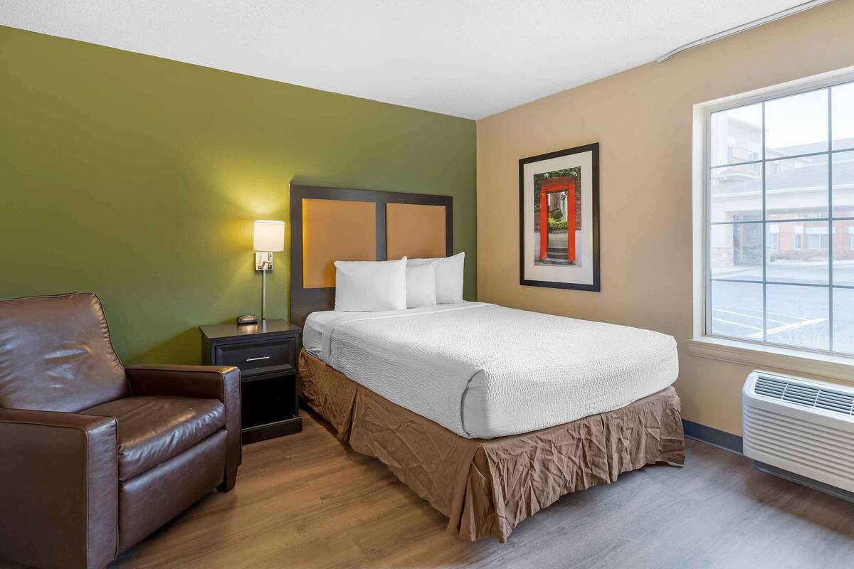 Une chambre d’hôtel avec un lit, un fauteuil en cuir marron, une table de chevet avec une lampe et un tableau encadré au mur.