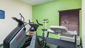 Sala de ginástica com equipamentos de exercício, uma balança fixada na parede e uma janela com persianas.