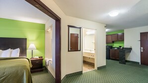 A hotel room with a bed, a nightstand, a lamp, a mirror, a bathroom with a sink and mirror, and a kitchenette with cabinets and a countertop. - Quality Inn & Suites Harvey - Chicago Southland (Harvey)