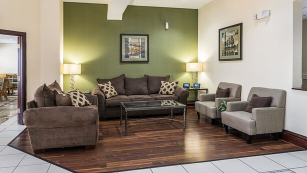 A living room with a brown sofa, a glass coffee table, and two armchairs. There are two wall-mounted lamps and framed pictures on the wall. - Quality Inn & Suites Harvey - Chicago Southland (Harvey)