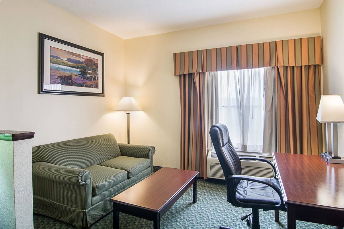 A hotel room with a green sofa, a wooden table, a black office chair, a framed picture on the wall, and a window with striped curtains.