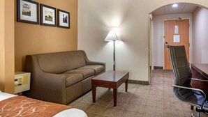 A hotel room with a bed, a brown sofa, a small wooden table, a desk with a chair, a lamp, and framed pictures on the wall. - Comfort Suites Hagerstown Southeast (Hagerstown)