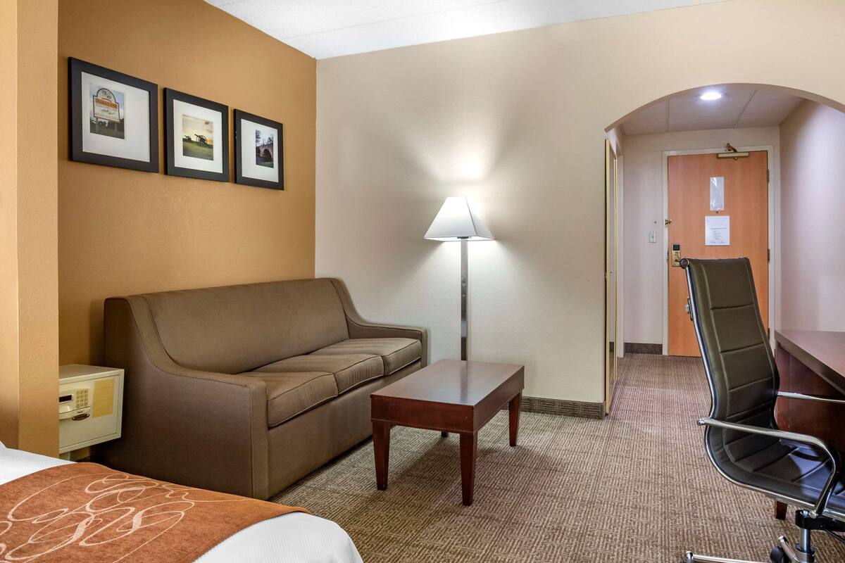A hotel room with a bed, a brown sofa, a small wooden table, a desk with a chair, a lamp, and framed pictures on the wall.