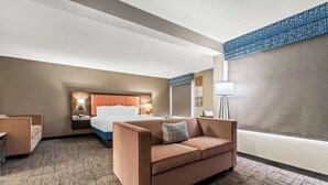 Ein Hotelzimmer mit braunem Sofa, einem Bett mit Kopfteil, einem Nachttisch, einer Lampe und Blick auf die Stadt.