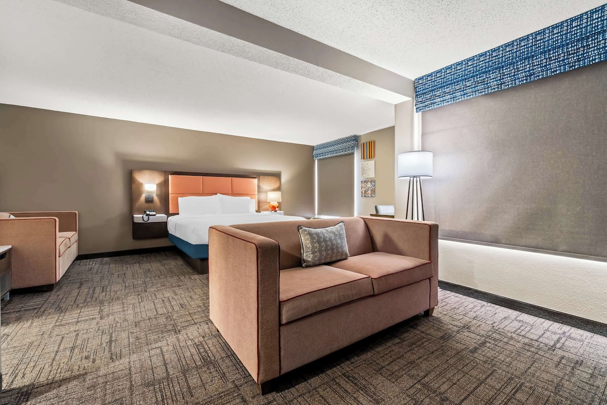 A hotel room with a brown sofa, a bed with a headboard, a nightstand, a lamp, and a view of the city.