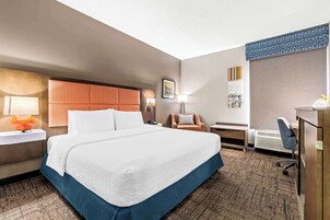 A hotel room with a large bed, a desk, a chair, a lamp, and a wall-mounted heater.