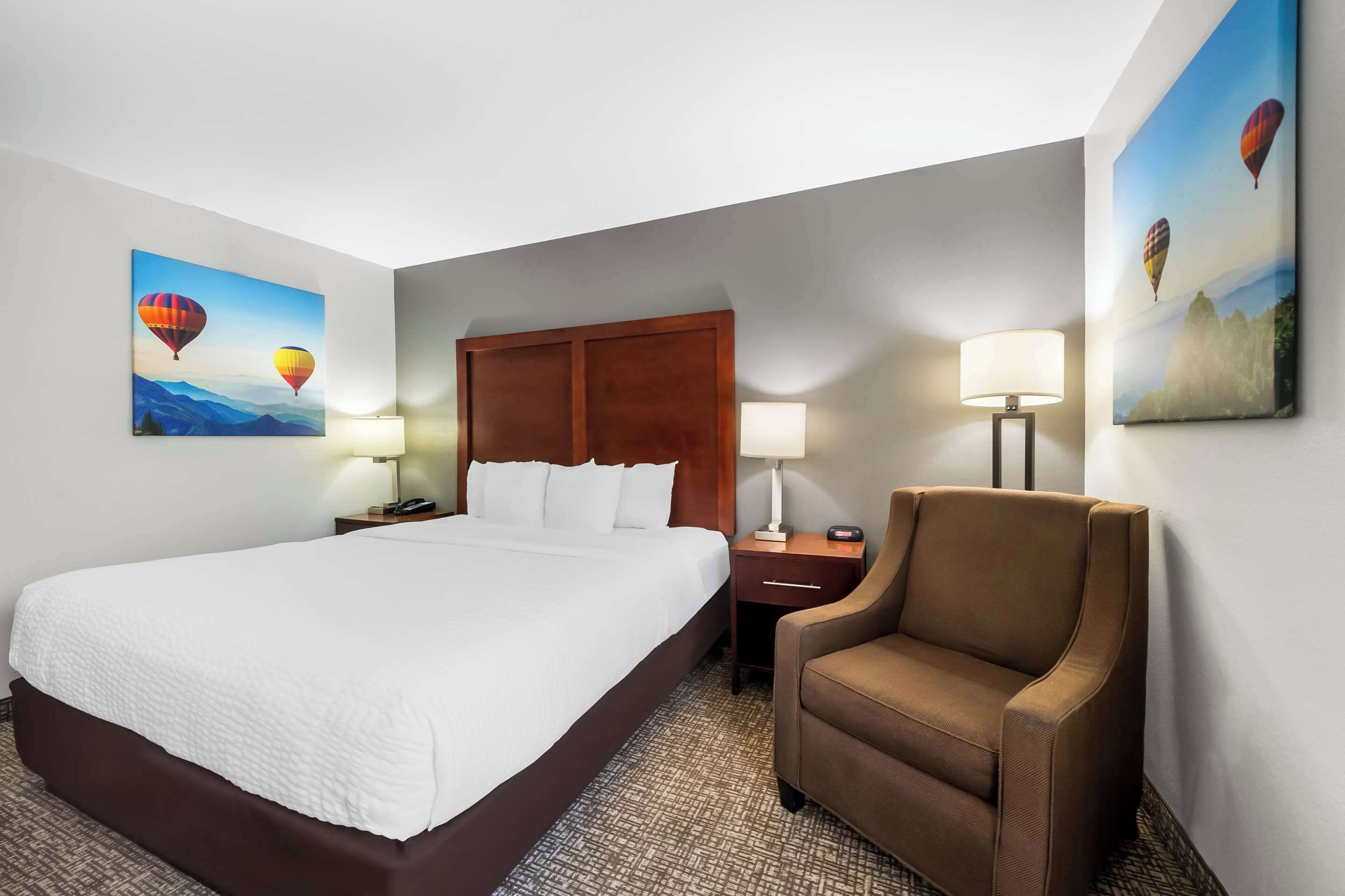 A hotel room with a large bed, a wooden headboard, two bedside tables with lamps, a brown armchair, and two wall-mounted paintings of hot air balloons.
