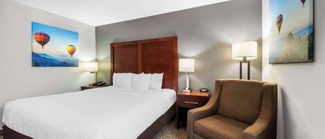 A hotel room with a large bed, a wooden headboard, two bedside tables with lamps, a brown armchair, and two wall-mounted paintings of hot air balloons.