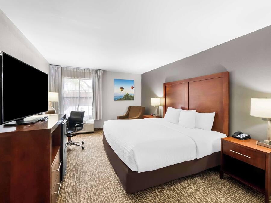 A hotel room with a large bed, a desk with a flat-screen TV, a chair, a desk lamp, and a nightstand with a lamp.
