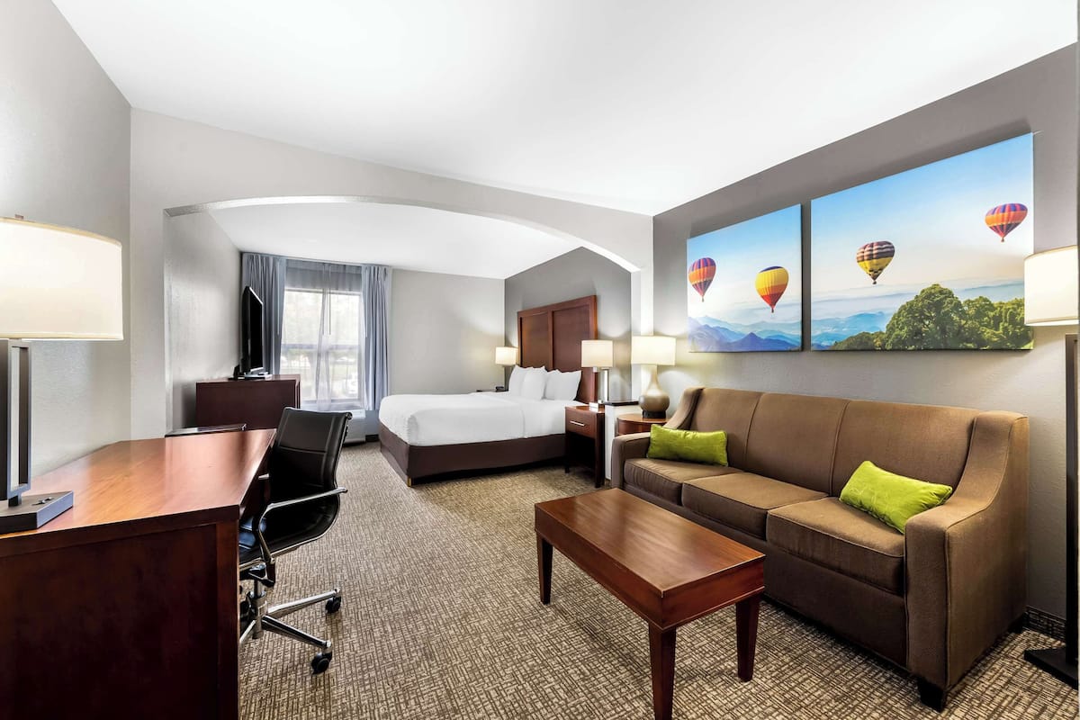 Une chambre d’hôtel avec un lit, un canapé, un bureau et une télévision. Il y a trois tableaux au mur et une fenêtre avec des rideaux.