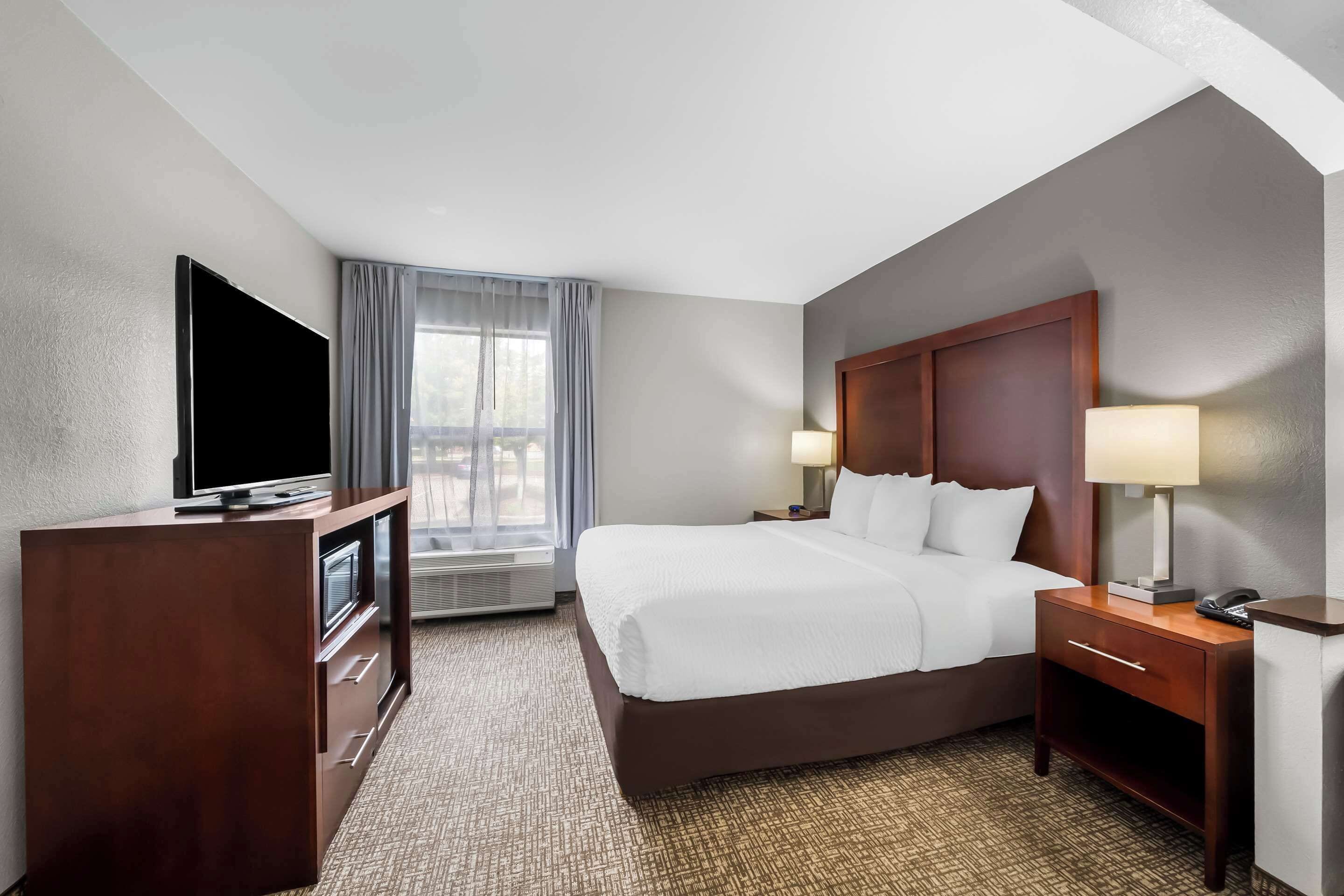 A hotel room with a large bed, a TV, a desk with a lamp, and a window with curtains.