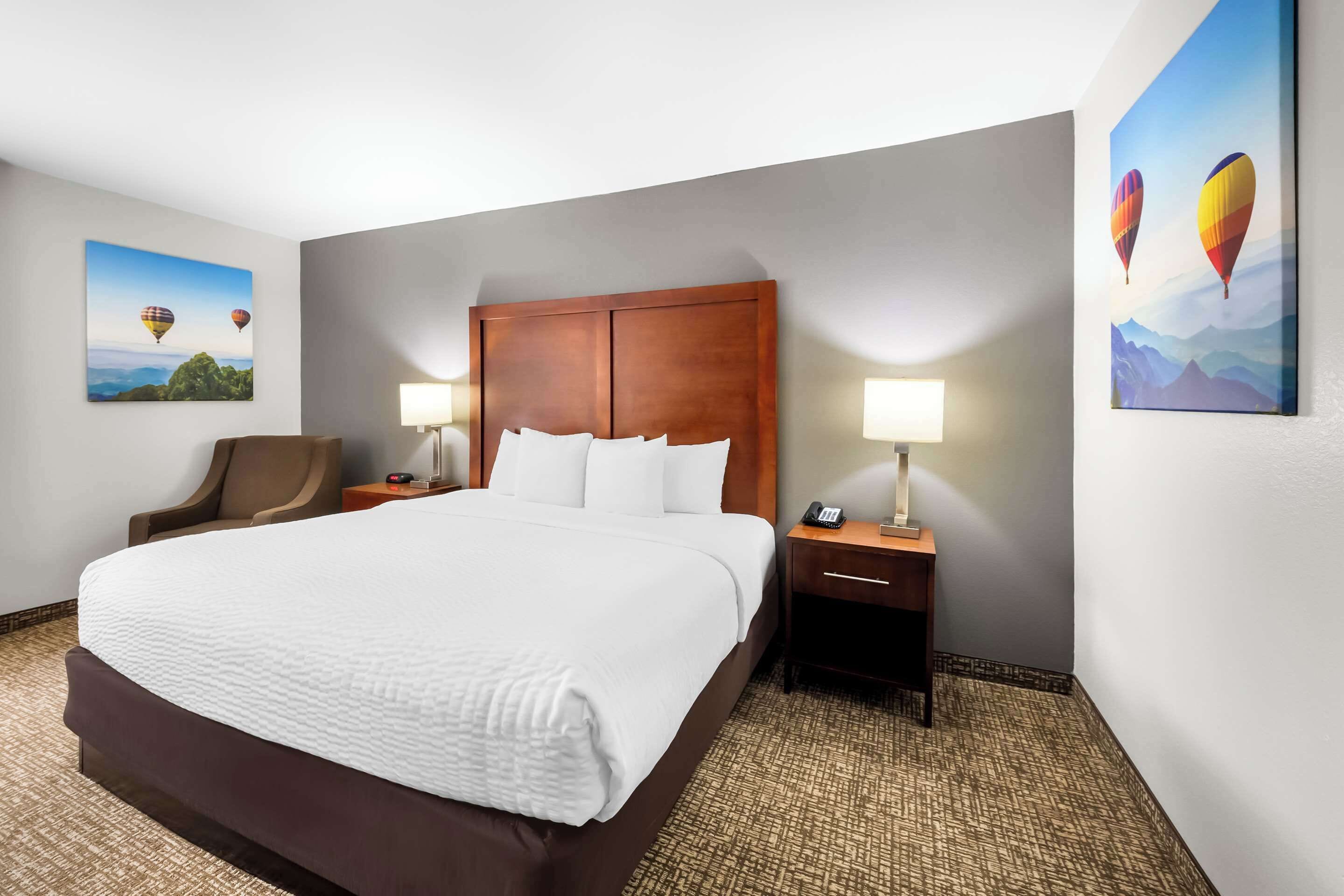 Habitación de hotel con una cama grande, dos mesitas de noche con lámparas, una silla y dos cuadros en las paredes.