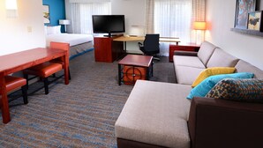 Quarto de hotel com cama, escrivaninha com cadeira, sofá, televisão e abajur.