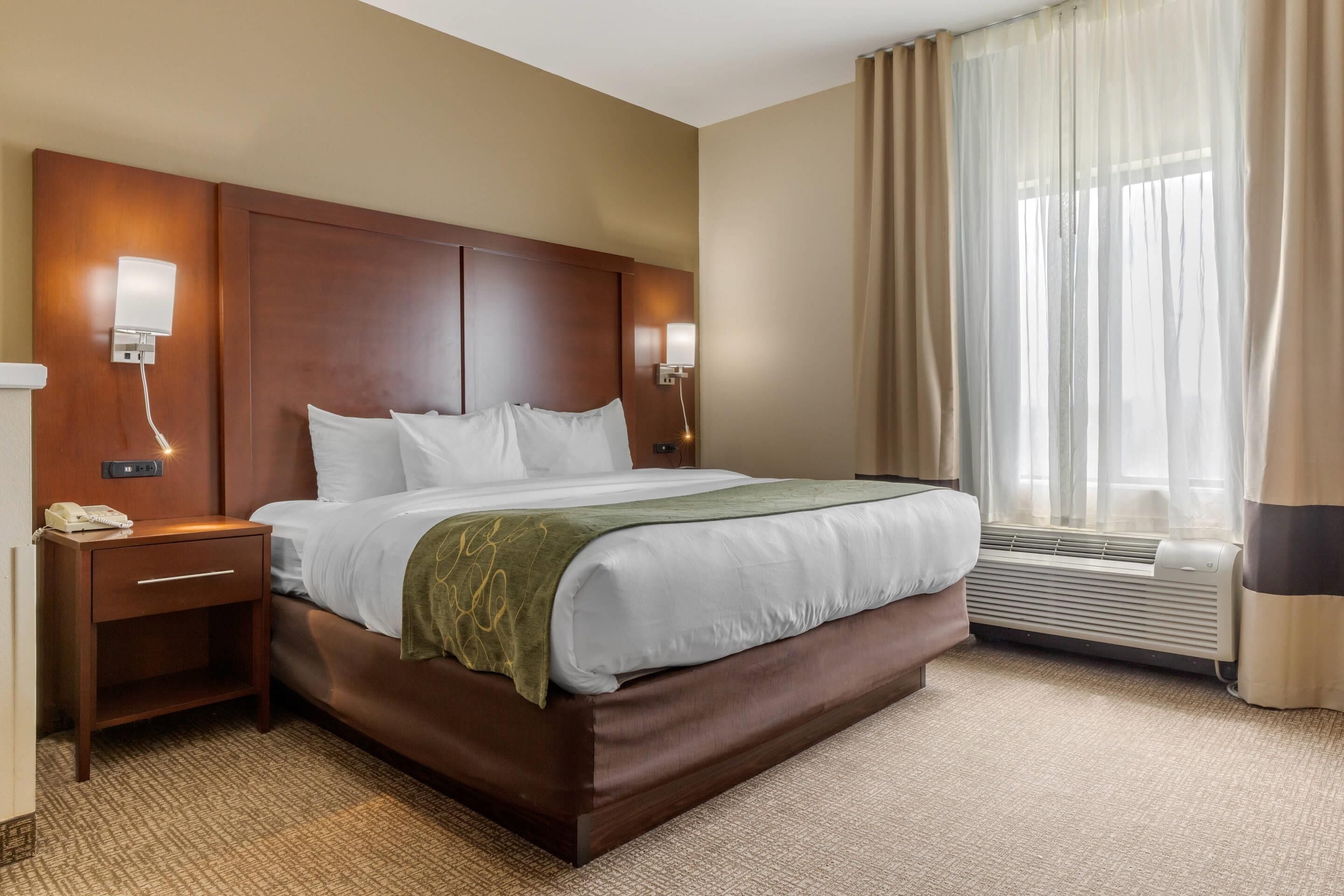 Ein Hotelzimmer mit einem großen Bett, Nachttischlampen, einem Telefon, einem fensterbelaubten Fenster und einer Klimaanlage.