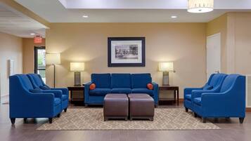 A lobby with a blue sofa set, two side chairs, a brown ottoman, a framed picture on the wall, and a patterned rug.